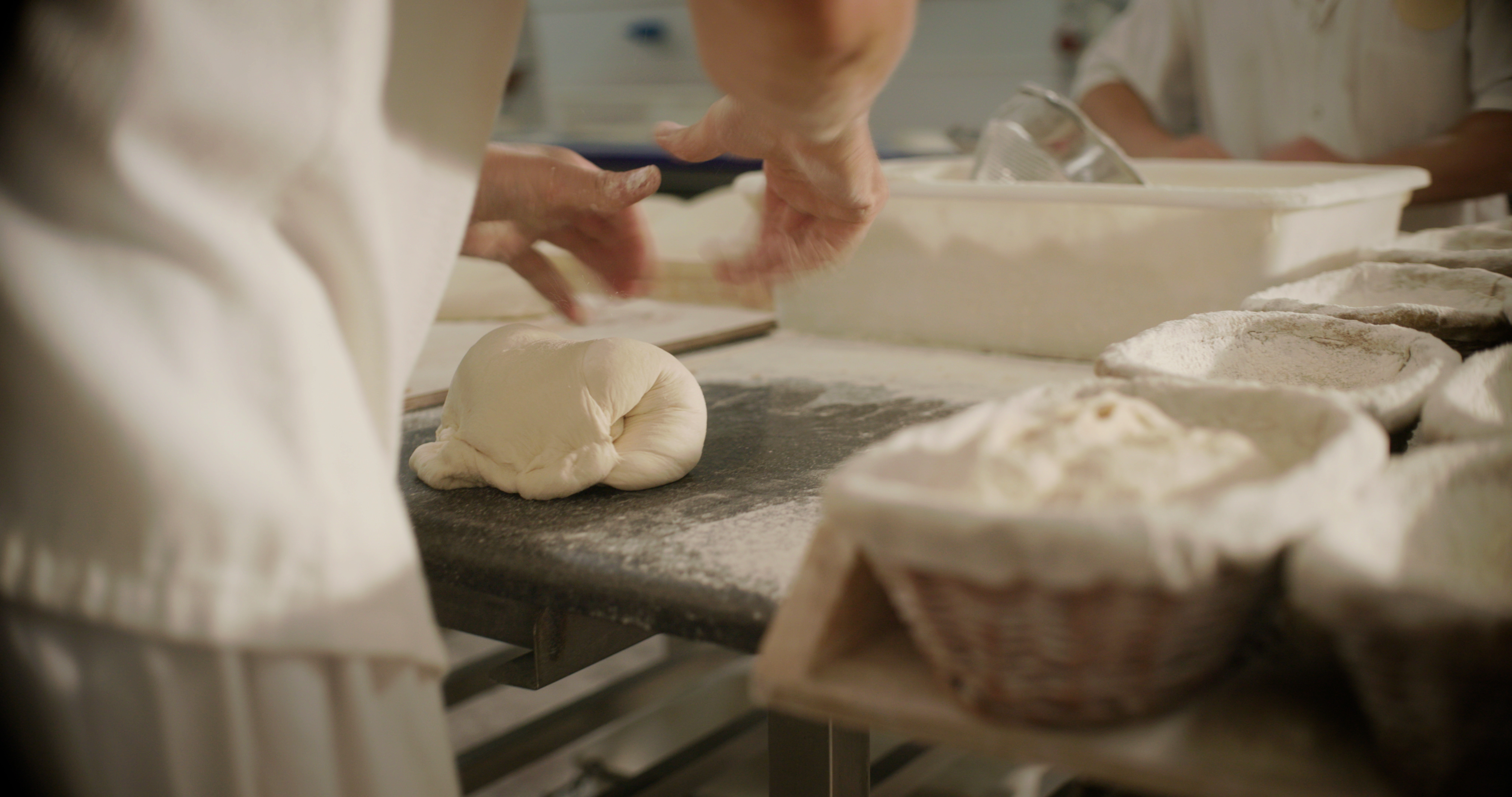 Baker preparing dough on a floured surface, baskets of dough in the background.