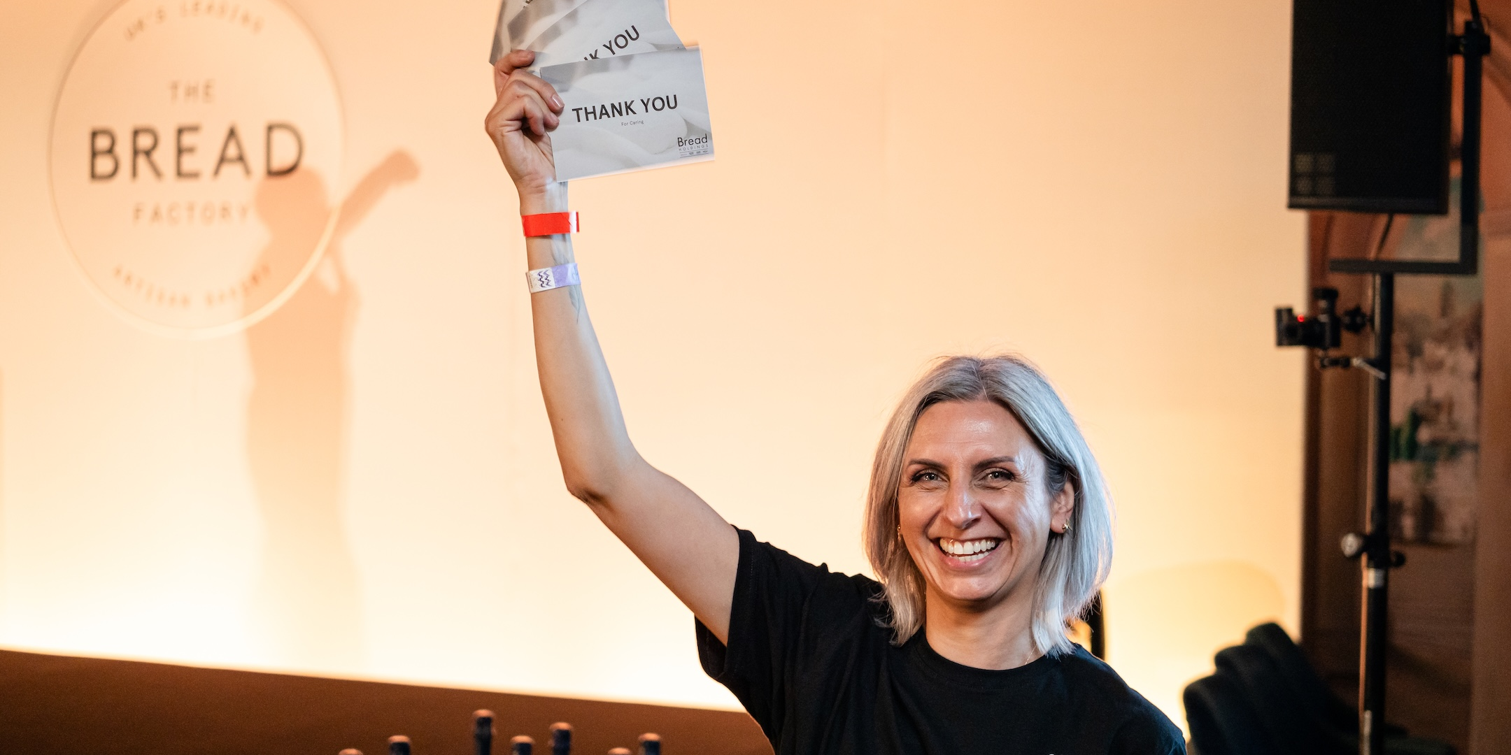 Woman holding "Thank You" cards, smiling, with wristbands, and "The Bread Factory" in the background.
