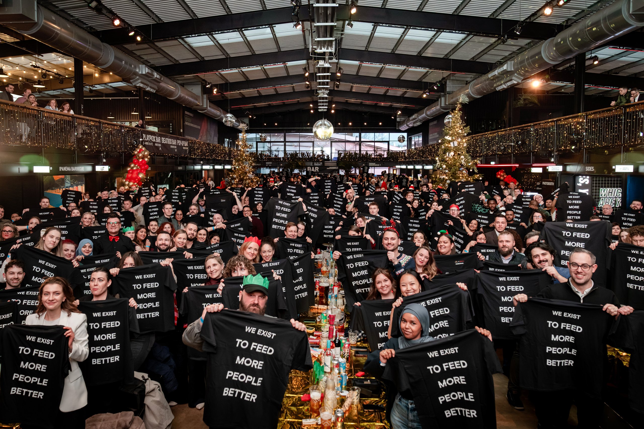 A large group of people hold up black t-shirts with the text "WE EXIST TO FEED MORE PEOPLE BETTER". A Christmas tree is visible in the background.