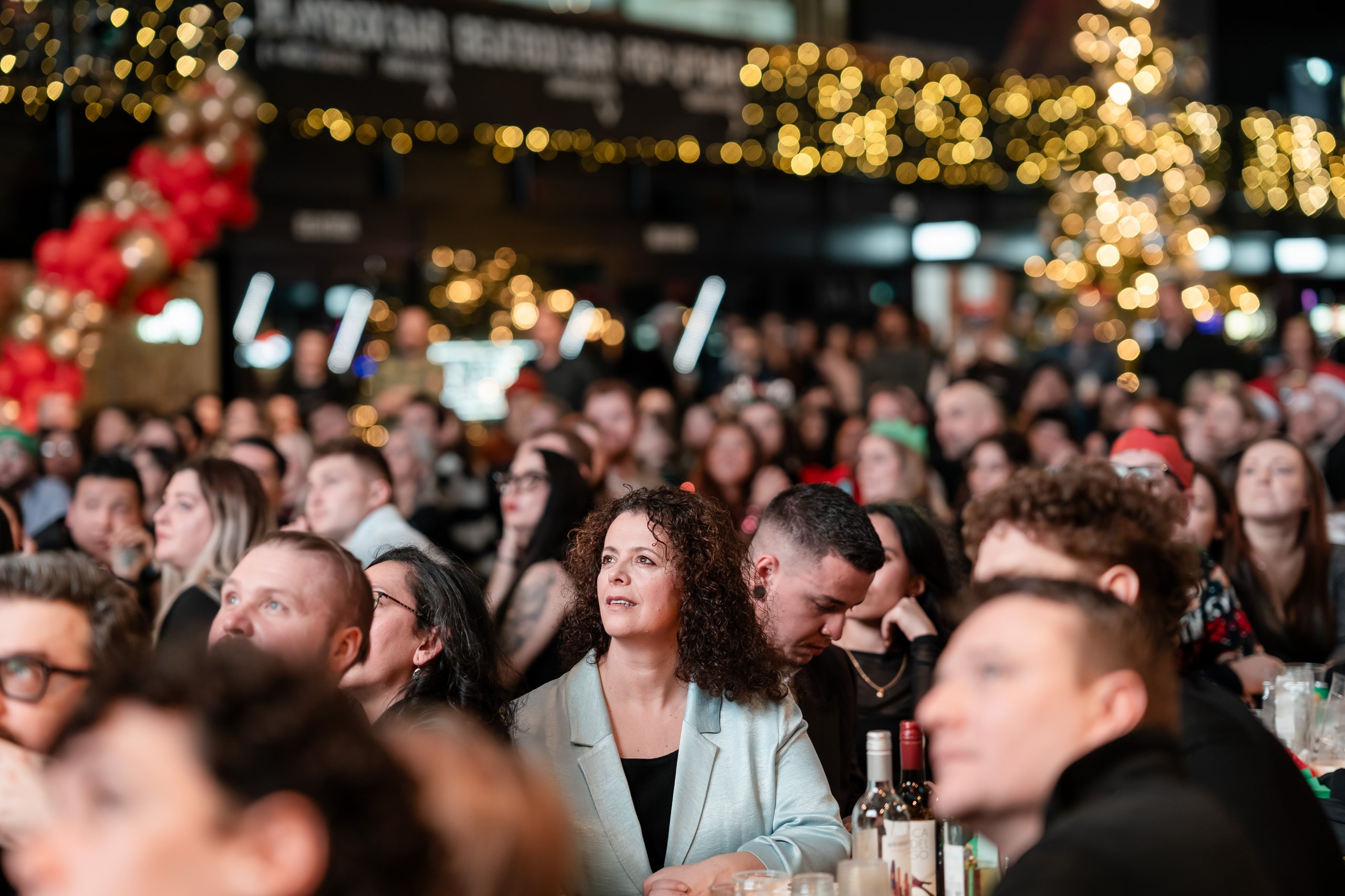 A crowd watches a performance at a festive event with blurred Christmas lights and decorations in the background.