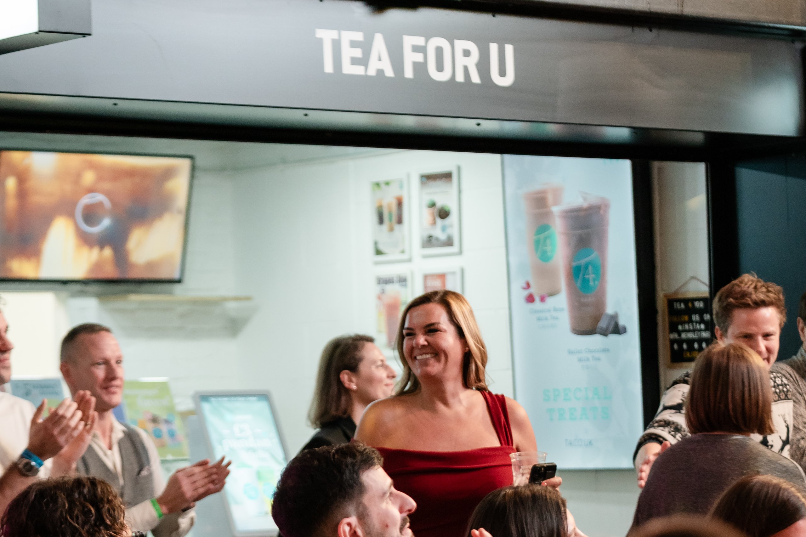 A crowd of people are clapping in an indoor venue. A sign above them reads "TEA FOR U". Some attendees are wearing wristbands.