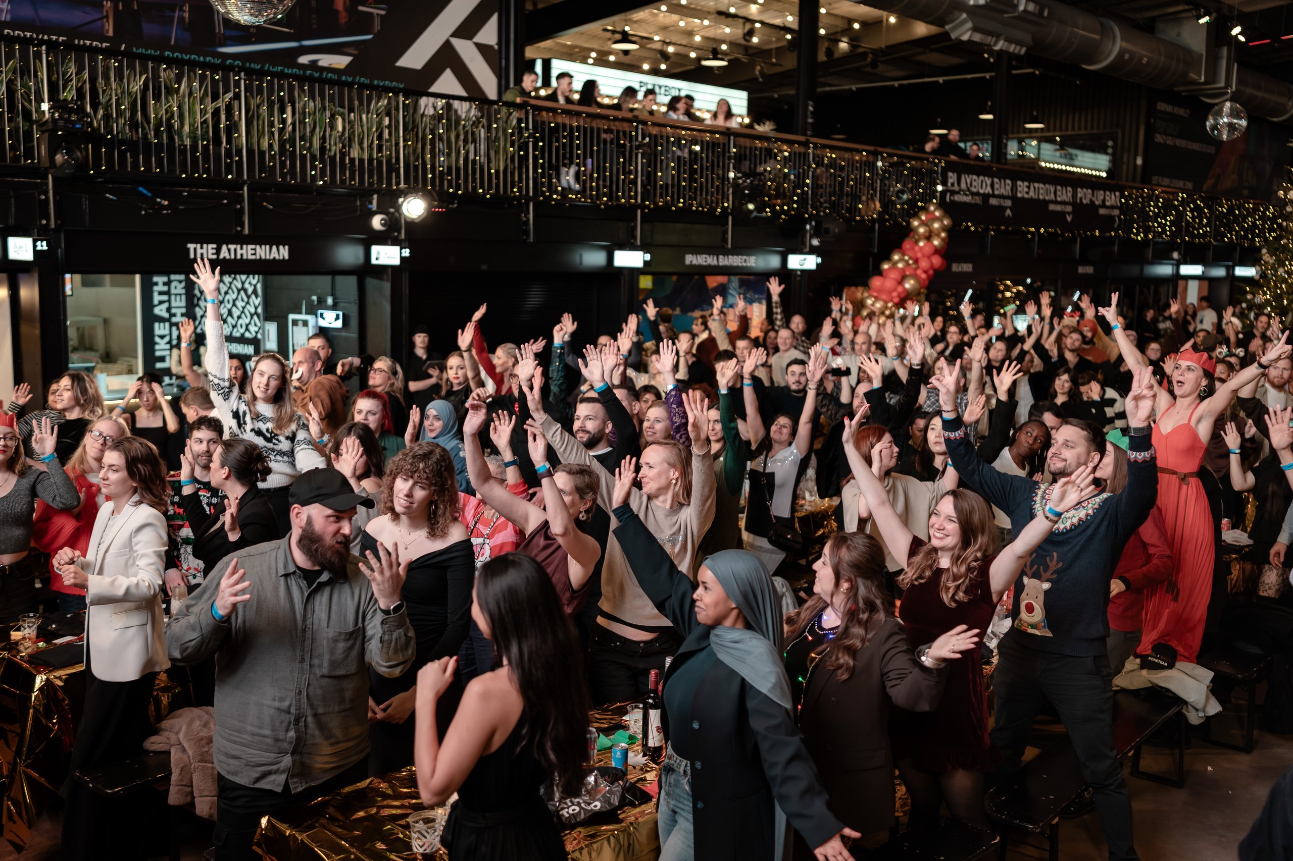 A diverse group of people are raising their hands in a lively party atmosphere with decorations and lights.