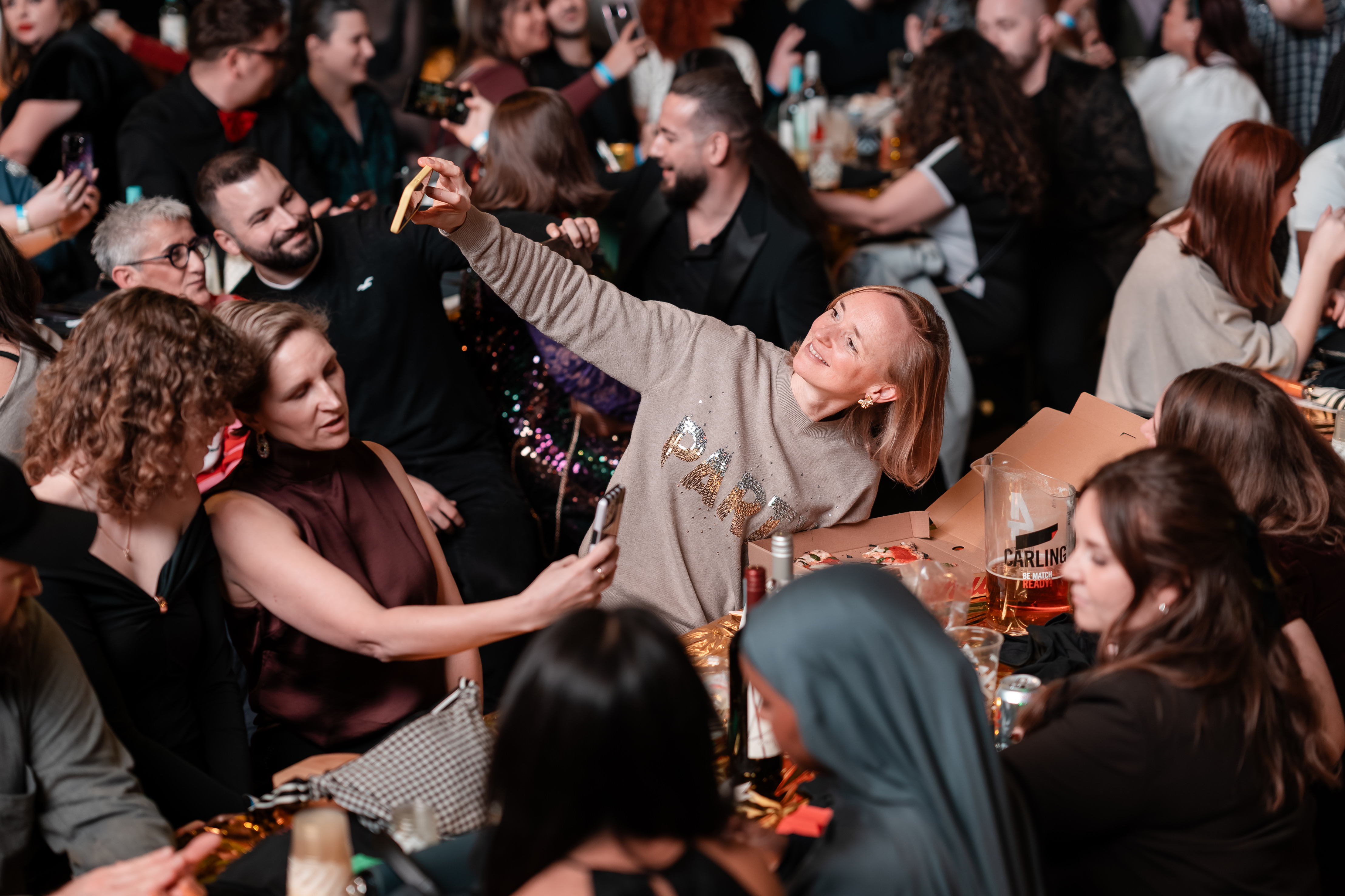 A woman in a sparkly sweater takes a selfie with her phone while sitting at a crowded table at a party.