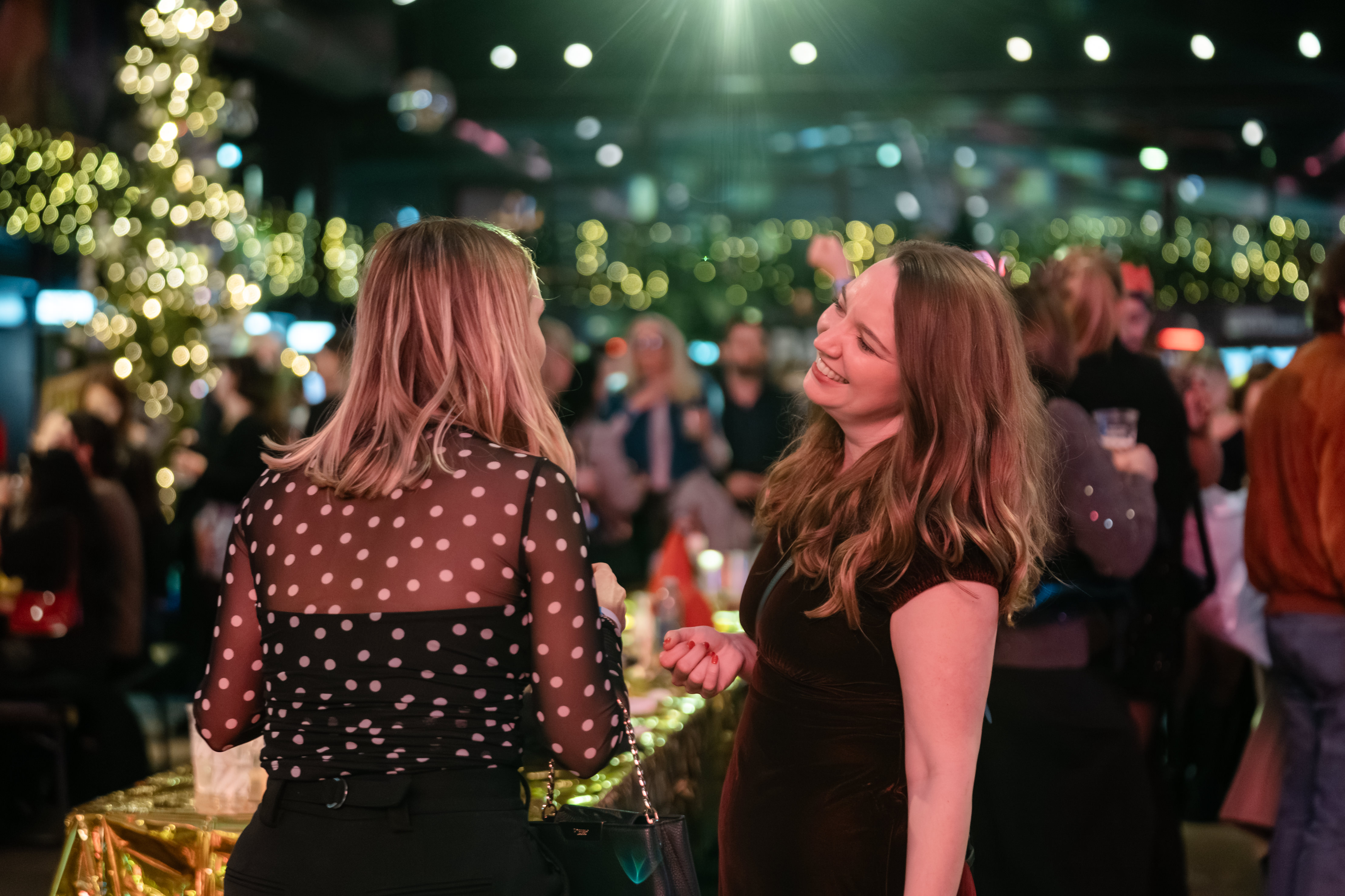 Two women laugh together at a festive holiday gathering with blurred bokeh lights in the background.
