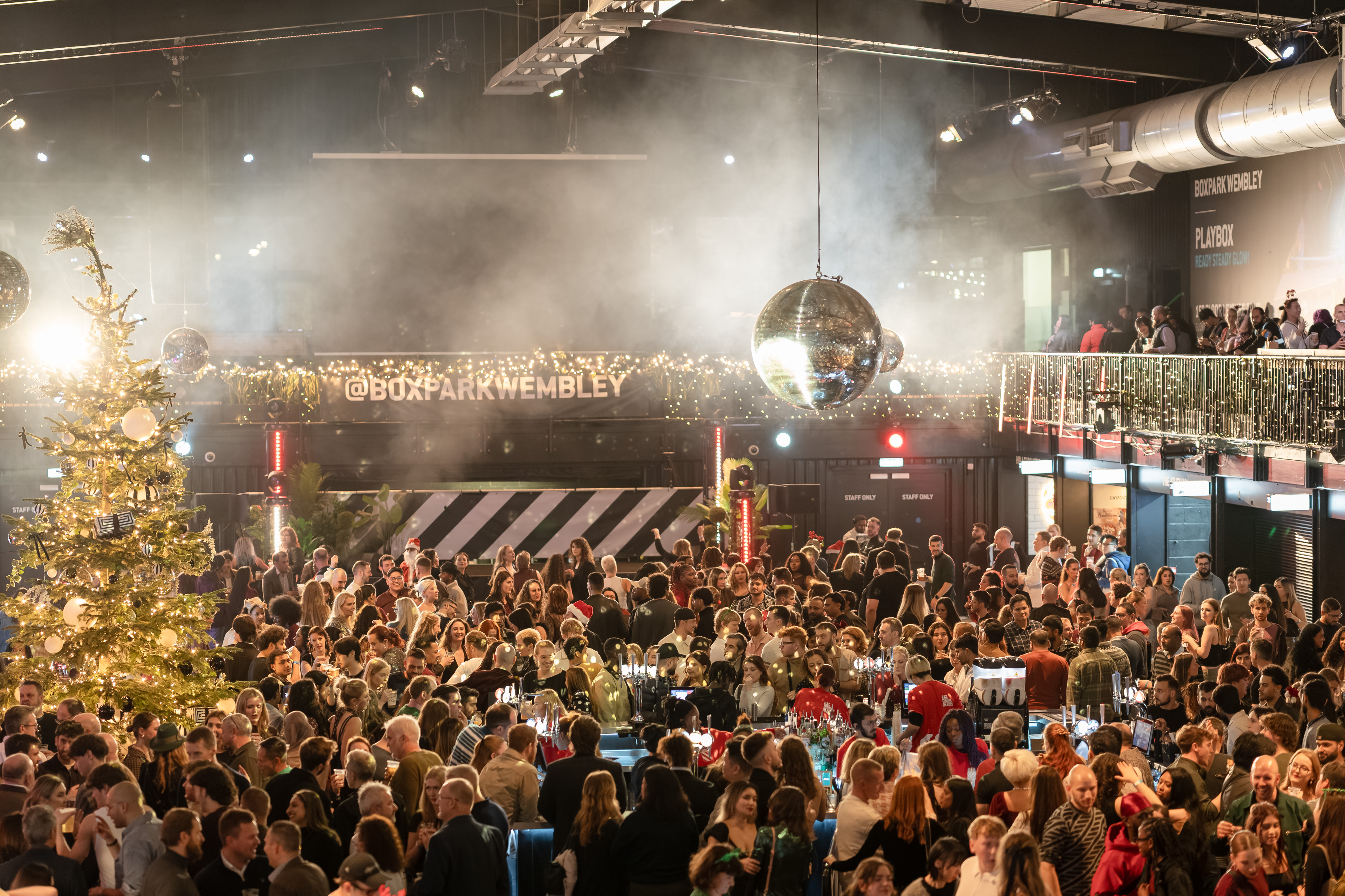 A crowd gathers under disco balls and a decorated Christmas tree at Boxpark Wembley, with fog and string lights creating a festive atmosphere.