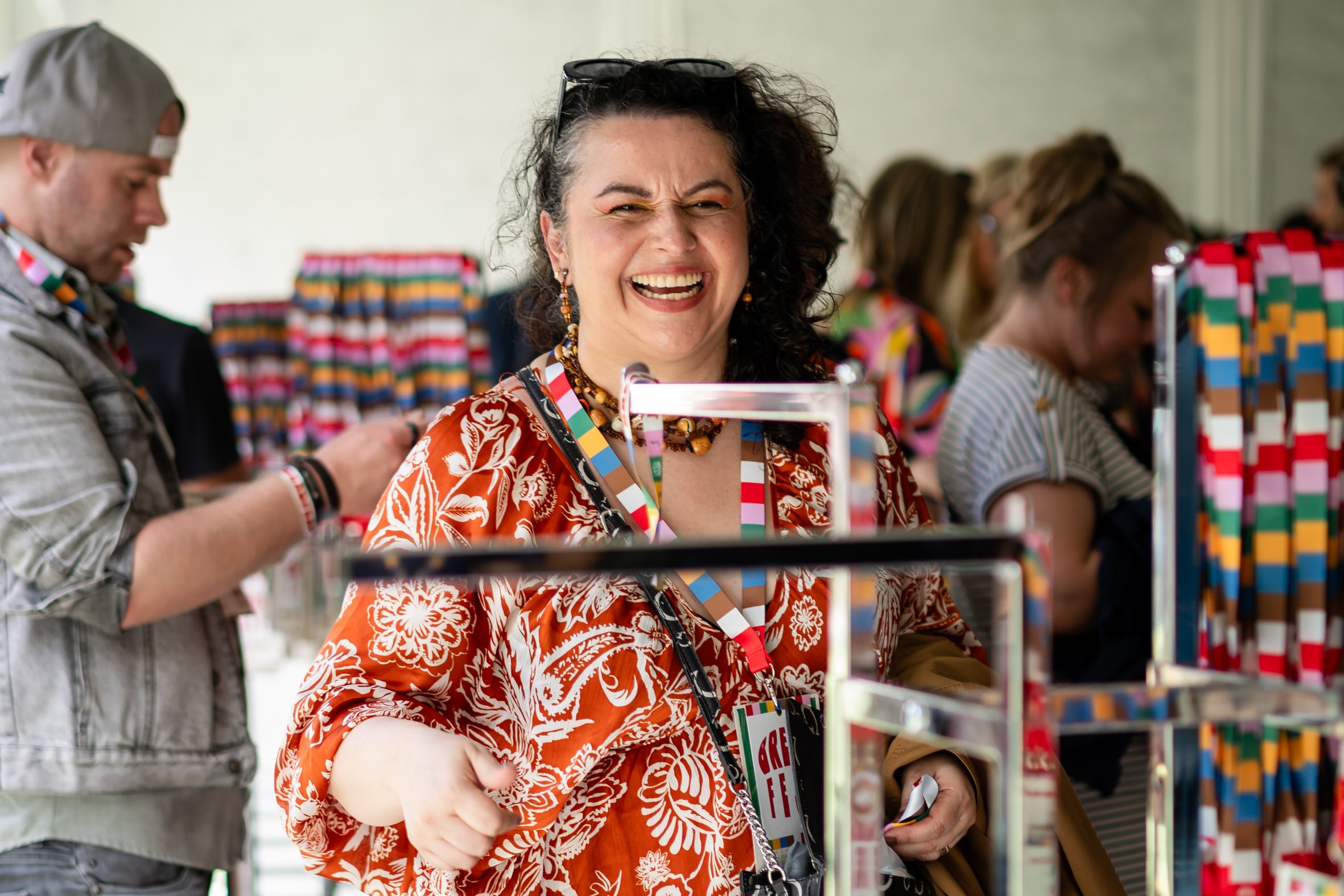 Woman laughs in an orange and white floral dress, wearing sunglasses on her head and multiple necklaces.