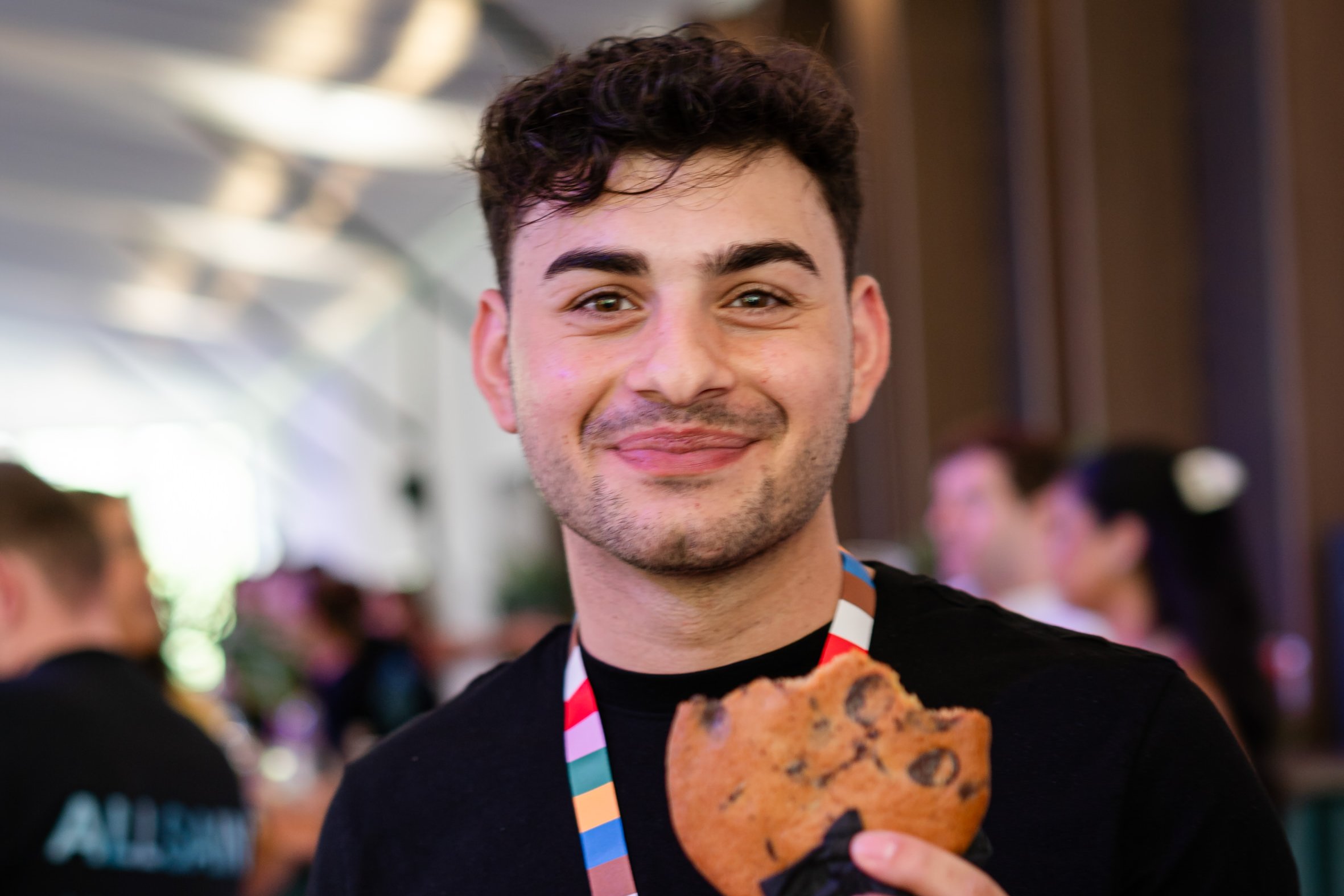 A smiling man wears a black t-shirt, a colorful lanyard, and a VIP access pass. He holds a large chocolate chip cookie.
