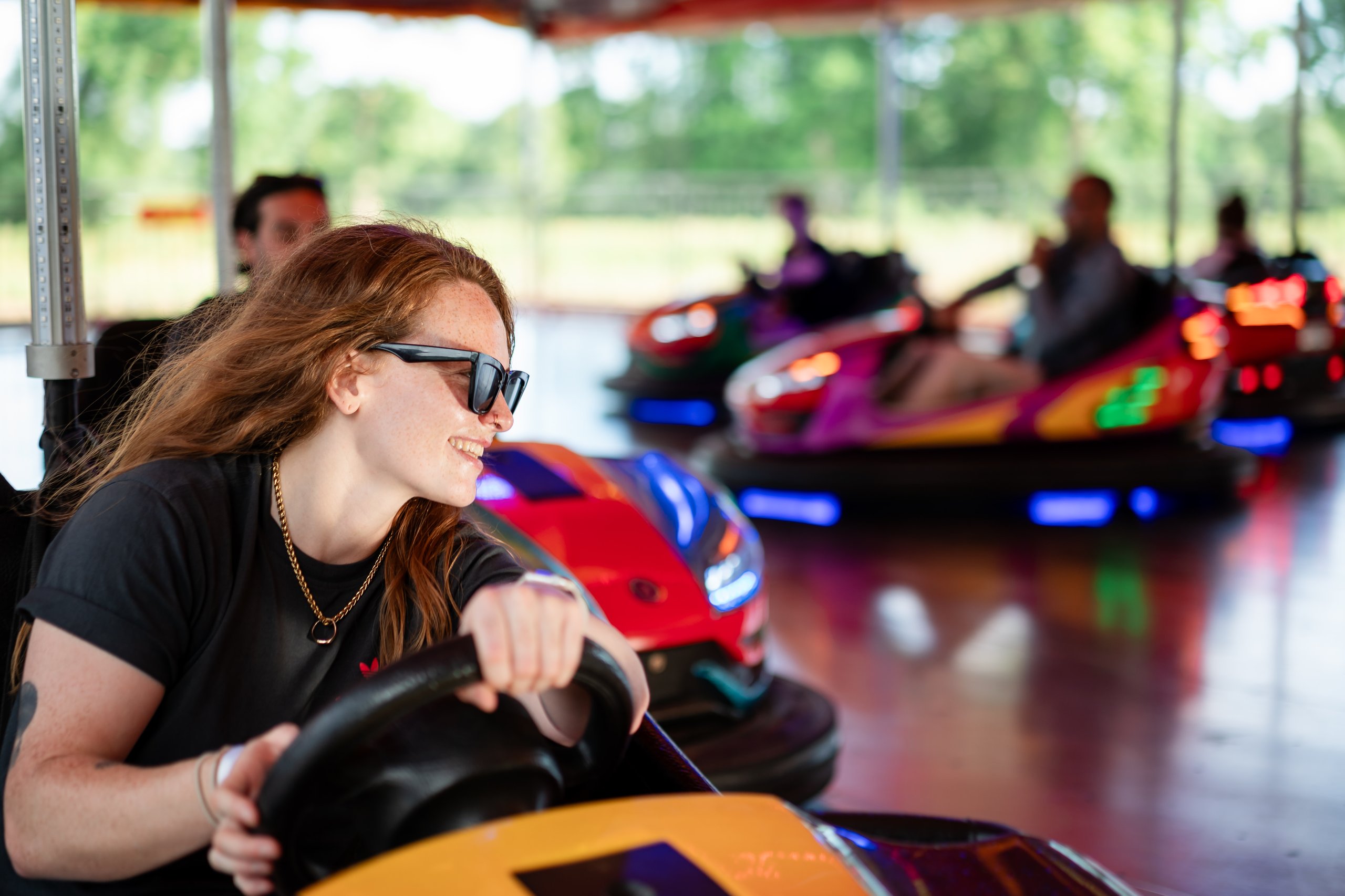 A young woman with freckles and sunglasses smiles as she drives a red bumper car at an amusement park.