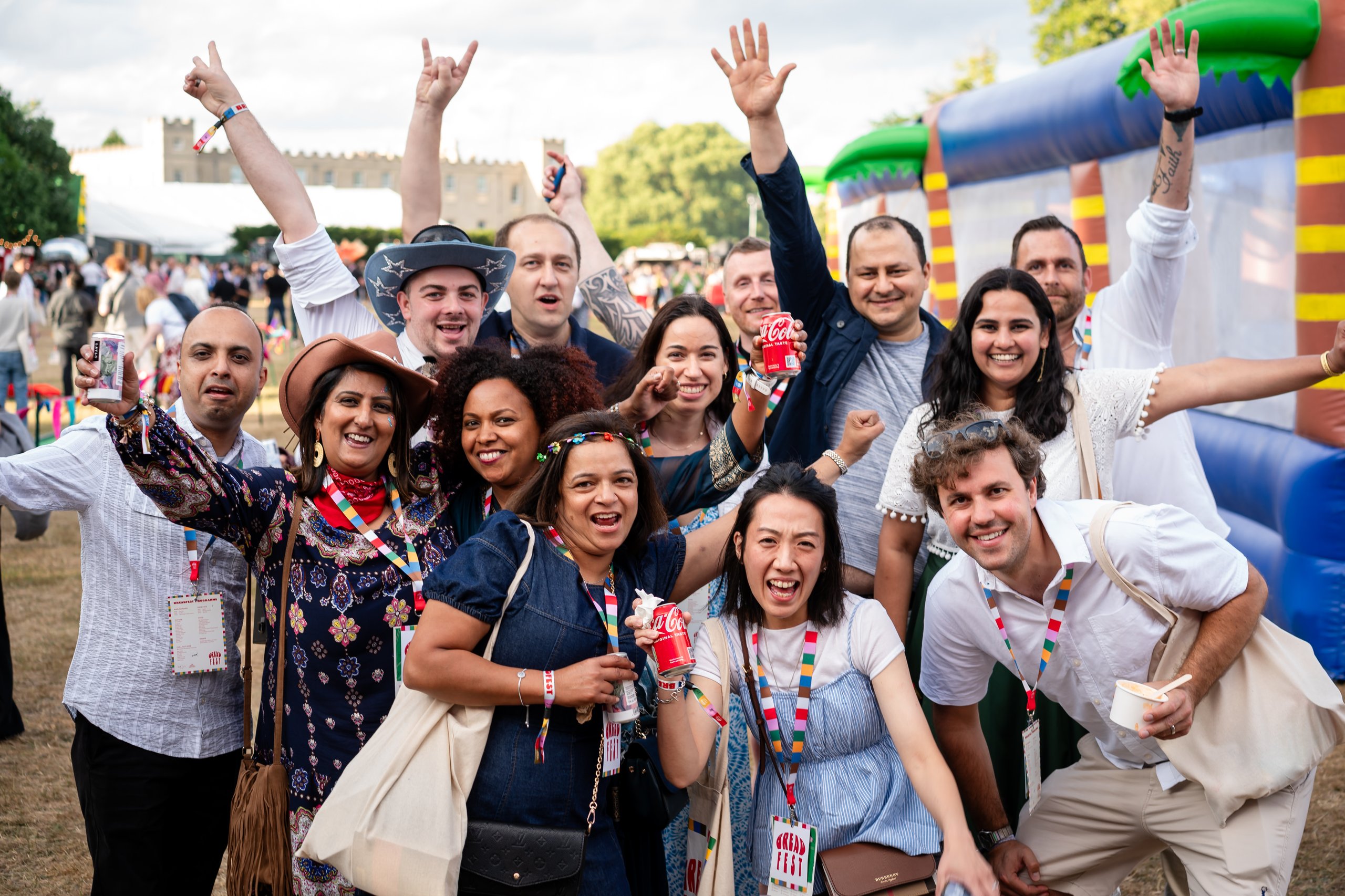 A diverse group of friends pose for a photo at an outdoor festival, smiling and holding drinks, with colorful banners and inflatable structures in the background.
